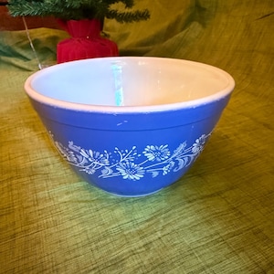 May include: A blue mixing bowl with a white floral pattern around the exterior. The bowl has a white interior and a slightly worn rim. A small Christmas tree is visible in the background.