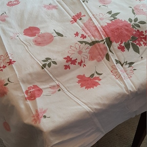 May include: White tablecloth with a repeating pattern of pink and red flowers and green leaves. The tablecloth is draped over a wooden table, with the legs of the table visible.