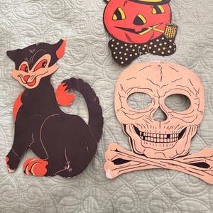 May include: Three vintage-style cardboard Halloween decorations: a black cat with orange accents, a pumpkin character wearing a black bowler hat and polka-dot bow tie while smoking a pipe, and a beige skull with crossbones featuring the text MADE IN U.S.A.