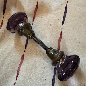 May include: A vintage door knob with a dark purple, faceted glass design. The knob has a brass-colored metal base and a long, threaded rod. The knob is set against a neutral-toned fabric with vertical purple and maroon stripes.