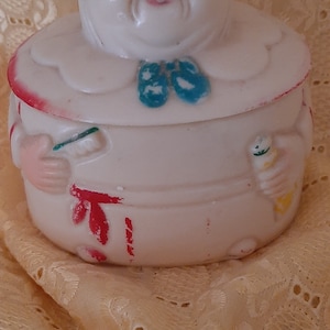 Puede incluir: Un tarro de galletas de cerámica blanca vintage con forma de anciana sonriente con gafas. El tarro tiene un lazo azul, detalles en rojo y está sobre una superficie texturizada.