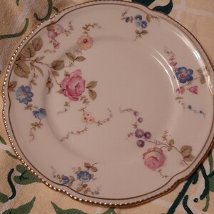 May include: A white porcelain plate with a scalloped edge and gold trim. The plate is decorated with pink, blue, and yellow floral patterns. The plate is set on a yellow and green patterned tablecloth.