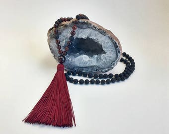 May include: A mala or prayer bead necklace with a burgundy tassel. The necklace is made of black lava rock beads and red carnelian beads. The necklace is displayed on a white agate geode.