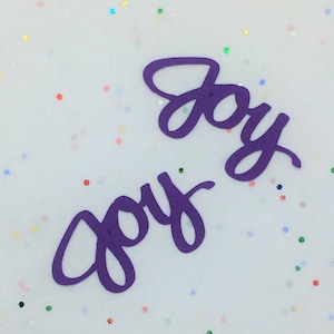 May include: Two purple paper cutouts of the word "joy" on a white background with colorful glitter.