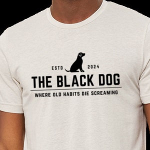 May include: A white t-shirt with a black graphic design featuring a black dog silhouette and the text "The Black Dog" with the tagline "Where old habits die screaming".