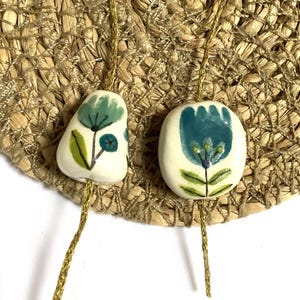 Pode incluir: Duas contas de cerâmica em branco sujo com desenhos florais pintados à mão em tons de azul-petróleo, azul e verde, cada uma enfiada num fio dourado fino e brilhante. A conta da esquerda é irregular, enquanto a da direita é redonda e lisa.