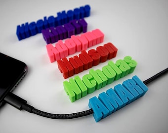 Pode incluir: Uma série de seis protetores de cabo de plástico impressos em 3D nas cores azul, roxo, rosa, vermelho, verde claro e ciano. Cada protetor apresenta um nome em relevo: Nicole-Marie, Kinley, Adelynn, Hudson, Nicholas e Amari, num cabo preto.
