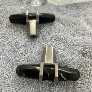 May include: Two black and silver cabinet knobs. Each knob has a black, oval-shaped handle with a silver metal base and brackets. The knobs are on a textured, light blue surface.