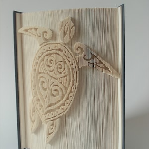 May include: A book art sculpture of a turtle with intricate patterns on its shell. The sculpture is made by folding the pages of a book into a three-dimensional shape.