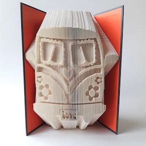 May include: A book folded into the shape of a vintage Volkswagen van with the word "love" written on the front. The book is open and the pages are folded to create the shape of the van. The book is on a white surface.