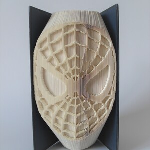 May include: A book folded into the shape of a Spiderman mask. The mask is white and has a black web design. The book is standing on a black surface.