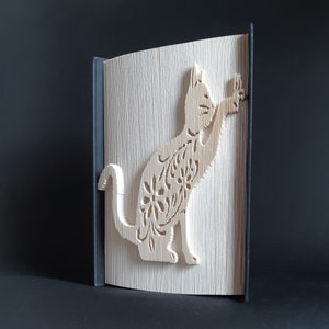 Könnte beinhalten: Ein weißes Buch mit einem 3D-Pop-up-Design einer Katze mit Blumenmustern. Die Katze sitzt mit dem Schwanz um den Körper gewickelt.