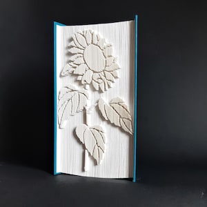 Op de afbeelding: Een wit papieren boek met een teal omslag, gevouwen tot een 3D-beeldhouwwerk van een zonnebloem.