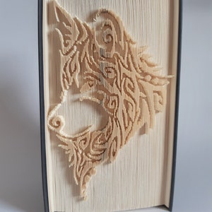 Könnte beinhalten: Ein Buch mit einem Wolfskopf, der in die Seiten geschnitten ist. Das Wolfsgesicht besteht aus gefalteten Seiten und ist hellbraun. Das Buch ist in Schwarz gebunden.