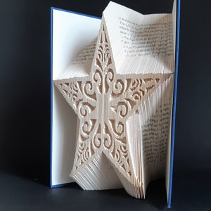 Op de afbeelding: Een boek met pagina's die zijn gevouwen en gesneden om een grote, sierlijke ster te vormen. Het ontwerp van de ster heeft ingewikkelde patronen. Het boek heeft een blauwe rug en de pagina's zijn lichtbeige van kleur. De achtergrond is zwart.