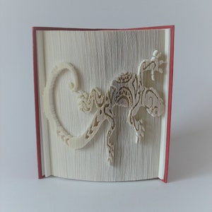Op de afbeelding: Een boek met een rode kaft is gevouwen in de vorm van een hagedis. De hagedis is wit en heeft een gedetailleerd ontwerp.