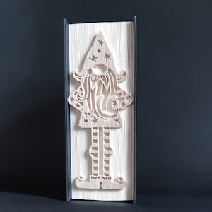 Peut inclure: Une sculpture de livre en papier blanc représentant un gnome portant un chapeau pointu avec des étoiles. Le gnome est debout, les bras tendus, et a une longue barbe.