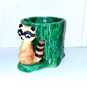 May include: A ceramic planter featuring a raccoon figure sitting next to a green tree stump. The raccoon has a black mask, brown fur, and a striped tail. The planter is green and has a hollow interior.