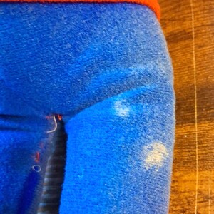 K&ouml;nnte beinhalten: Eine Pl&uuml;schpuppe mit rotem Oberteil und blauer Hose. Die Beine der Puppe sind blau, und der untere Teil der Beine ist gelb. Die Puppe befindet sich auf einer Holzoberfl&auml;che.