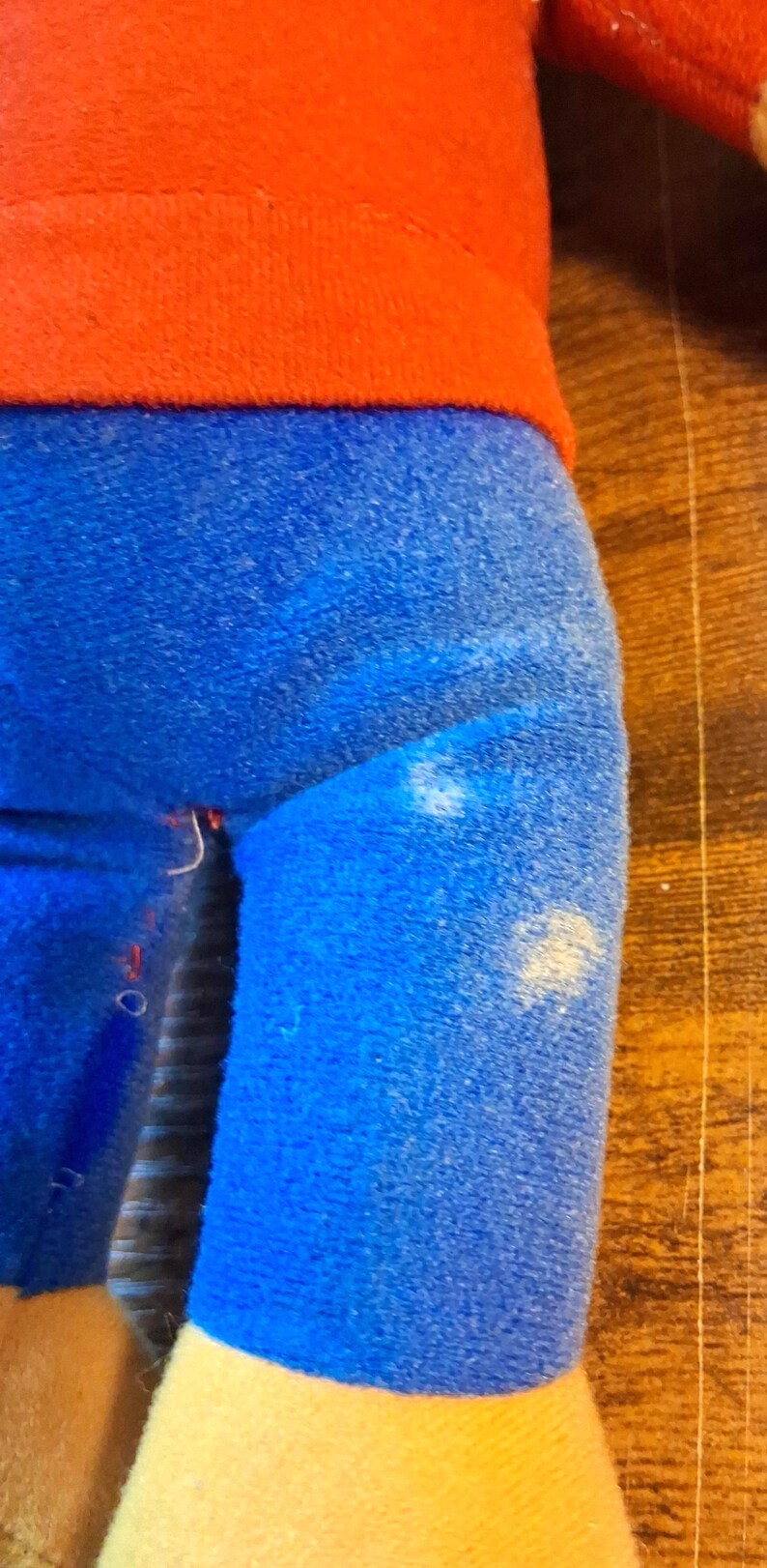 K&ouml;nnte beinhalten: Eine Pl&uuml;schpuppe mit rotem Oberteil und blauer Hose. Die Beine der Puppe sind blau, und der untere Teil der Beine ist gelb. Die Puppe befindet sich auf einer Holzoberfl&auml;che.