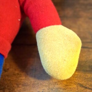 K&ouml;nnte beinhalten: Nahaufnahme von Arm und Fu&szlig; eines Pl&uuml;schtiers. Der Arm ist rot und geht in einen gelben Fu&szlig; &uuml;ber. Der Arm des Spielzeugs besteht aus einem weichen, flauschigen Material. Der Fu&szlig; ist hellgelb.