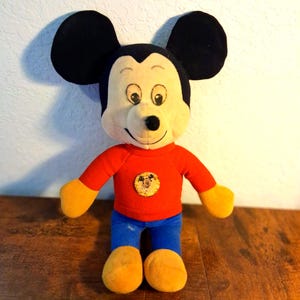 K&ouml;nnte beinhalten: Eine Mickey Mouse-Pl&uuml;schpuppe mit gro&szlig;en schwarzen Ohren, einem cremefarbenen Gesicht und einem roten Hemd. Die Puppe hat gelbe H&auml;nde und F&uuml;&szlig;e, eine blaue Hose und ein rundes Emblem auf dem Hemd. Die Puppe steht auf einer Holzoberfl&auml;che.