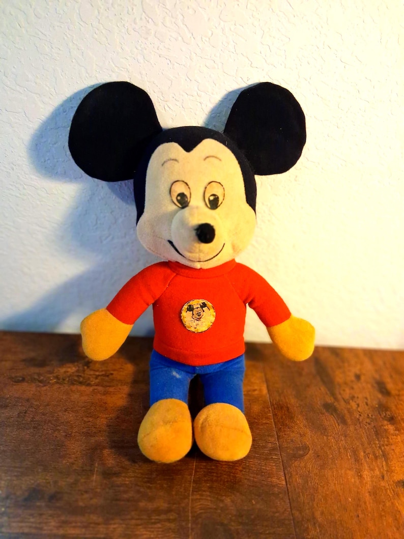 K&ouml;nnte beinhalten: Eine Mickey Mouse-Pl&uuml;schpuppe mit gro&szlig;en schwarzen Ohren, einem cremefarbenen Gesicht und einem roten Hemd. Die Puppe hat gelbe H&auml;nde und F&uuml;&szlig;e, eine blaue Hose und ein rundes Emblem auf dem Hemd. Die Puppe steht auf einer Holzoberfl&auml;che.