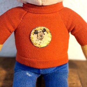 K&ouml;nnte beinhalten: Vintage Pl&uuml;schpuppe mit rotem Pullover, Mickey Mouse-Emblem, blauer Jeans und gelben H&auml;nden. Die Puppe hat einen wei&szlig;en Kopf, und das Design erinnert an klassische Zeichentrickfiguren.