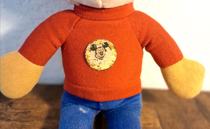 K&ouml;nnte beinhalten: Vintage Pl&uuml;schpuppe mit rotem Pullover, Mickey Mouse-Emblem, blauer Jeans und gelben H&auml;nden. Die Puppe hat einen wei&szlig;en Kopf, und das Design erinnert an klassische Zeichentrickfiguren.