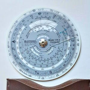 May include: A clear, circular Jeppesen Sanderson Model CR-2 Computer with multiple scales and markings for aviation calculations. The device features a central brass fitting and is designed for determining true airspeed, altitude, and other flight data.