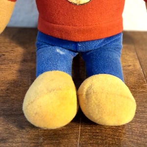 K&ouml;nnte beinhalten: Ein Stofftier mit rotem Oberteil, blauer Hose und gelben F&uuml;&szlig;en. Die Arme und F&uuml;&szlig;e des Spielzeugs sind hellgelb. Es befindet sich auf einer Holzoberfl&auml;che.