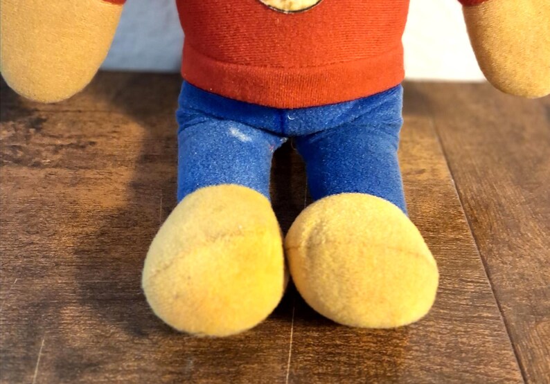 K&ouml;nnte beinhalten: Ein Stofftier mit rotem Oberteil, blauer Hose und gelben F&uuml;&szlig;en. Die Arme und F&uuml;&szlig;e des Spielzeugs sind hellgelb. Es befindet sich auf einer Holzoberfl&auml;che.