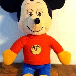 K&ouml;nnte beinhalten: Ein Vintage-Mickey-Mouse-Pl&uuml;schtier. Das Pl&uuml;schtier hat ein cremefarbenes Gesicht, schwarze Ohren und tr&auml;gt ein rotes Oberteil, eine blaue Hose sowie gelbe Handschuhe und Schuhe. Ein kreisf&ouml;rmiger Aufn&auml;her mit Mickys Gesicht befindet sich auf dem Oberteil.