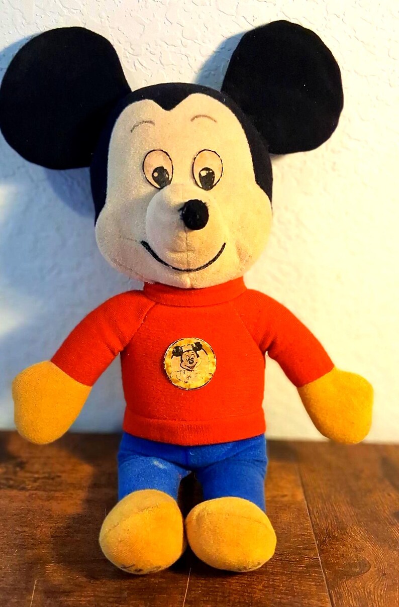 K&ouml;nnte beinhalten: Ein Vintage-Mickey-Mouse-Pl&uuml;schtier. Das Pl&uuml;schtier hat ein cremefarbenes Gesicht, schwarze Ohren und tr&auml;gt ein rotes Oberteil, eine blaue Hose sowie gelbe Handschuhe und Schuhe. Ein kreisf&ouml;rmiger Aufn&auml;her mit Mickys Gesicht befindet sich auf dem Oberteil.