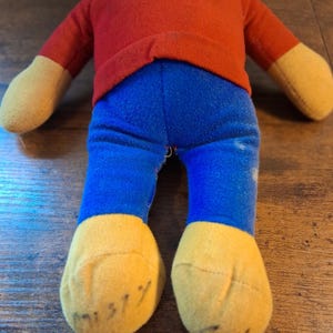 K&ouml;nnte beinhalten: Eine Vintage-Pl&uuml;schpuppe mit rotem Oberteil, blauer Hose und gelben H&auml;nden und F&uuml;&szlig;en. Die F&uuml;&szlig;e der Puppe haben handschriftliche Markierungen. Die Puppe ist aus weichem Stoff und scheint gut erhalten zu sein. Das Spielzeug ist wahrscheinlich ein Sammlerst&uuml;ck.