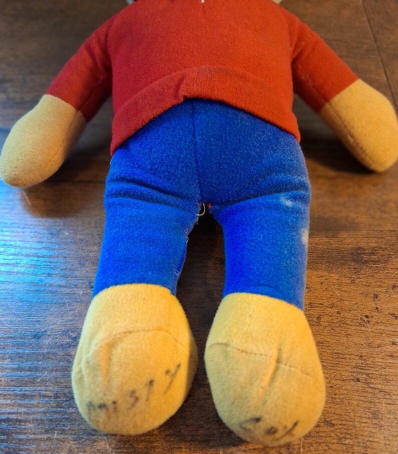 K&ouml;nnte beinhalten: Eine Vintage-Pl&uuml;schpuppe mit rotem Oberteil, blauer Hose und gelben H&auml;nden und F&uuml;&szlig;en. Die F&uuml;&szlig;e der Puppe haben handschriftliche Markierungen. Die Puppe ist aus weichem Stoff und scheint gut erhalten zu sein. Das Spielzeug ist wahrscheinlich ein Sammlerst&uuml;ck.