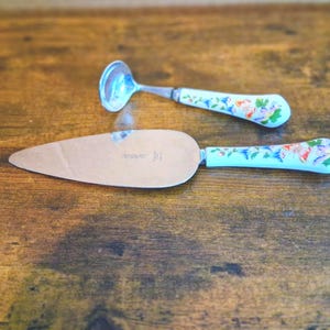 May include: A cake server and a small ladle with matching floral handles rest on a wooden surface. The handles are light blue with a colorful floral pattern. The cake server has a silver blade and the ladle has a silver bowl.