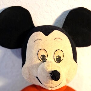 K&ouml;nnte beinhalten: Nahaufnahme eines Mickey Mouse Pl&uuml;schtiers. Das Spielzeug hat gro&szlig;e schwarze Ohren, ein cremefarbenes Gesicht mit schwarzen Augen, eine schwarze Nase und ein rotes Hemd. Das Pl&uuml;schtier ist eine klassische Darstellung der ikonischen Zeichentrickfigur.