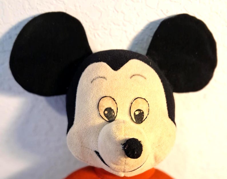 K&ouml;nnte beinhalten: Nahaufnahme eines Mickey Mouse Pl&uuml;schtiers. Das Spielzeug hat gro&szlig;e schwarze Ohren, ein cremefarbenes Gesicht mit schwarzen Augen, eine schwarze Nase und ein rotes Hemd. Das Pl&uuml;schtier ist eine klassische Darstellung der ikonischen Zeichentrickfigur.