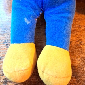 K&ouml;nnte beinhalten: Nahaufnahme der Beine eines Pl&uuml;schtiers. Die Beine sind blau mit gelben F&uuml;&szlig;en. Die Beine des Spielzeugs bestehen aus einem weichen, flauschigen Material. Auf dem linken Bein befindet sich ein Fleck.