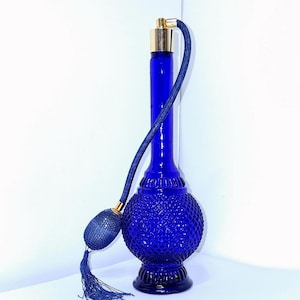 May include: A cobalt blue perfume bottle with a textured, diamond-patterned body and a tall, slender neck. It features a gold-colored atomizer and a matching blue atomizer bulb with a tassel. The bottle is approximately 8 inches tall.