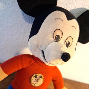K&ouml;nnte beinhalten: Ein Vintage-Mickey-Mouse-Pl&uuml;schtier. Das Pl&uuml;schtier hat ein wei&szlig;es Gesicht, schwarze Ohren und ein rotes Hemd mit einem kreisf&ouml;rmigen Aufn&auml;her. Das Spielzeug hat ein klassisches Design mit einem l&auml;chelnden Gesichtsausdruck und gro&szlig;en Augen. Das Pl&uuml;schtier ist ein nostalgisches Sammlerst&uuml;ck.