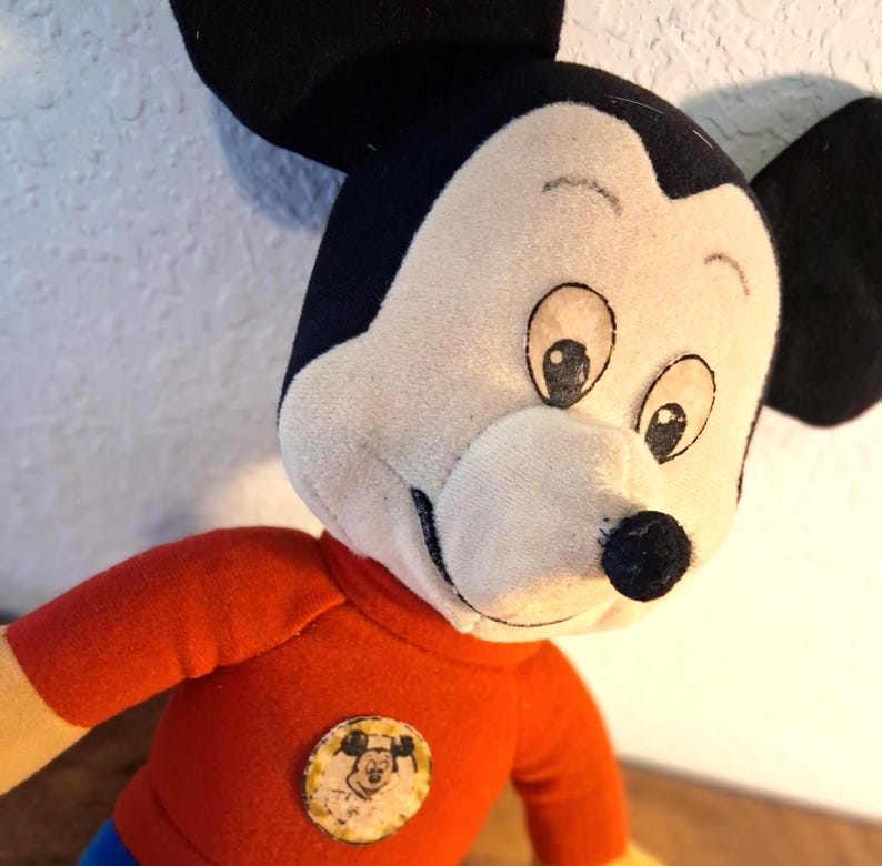 K&ouml;nnte beinhalten: Ein Vintage-Mickey-Mouse-Pl&uuml;schtier. Das Pl&uuml;schtier hat ein wei&szlig;es Gesicht, schwarze Ohren und ein rotes Hemd mit einem kreisf&ouml;rmigen Aufn&auml;her. Das Spielzeug hat ein klassisches Design mit einem l&auml;chelnden Gesichtsausdruck und gro&szlig;en Augen. Das Pl&uuml;schtier ist ein nostalgisches Sammlerst&uuml;ck.