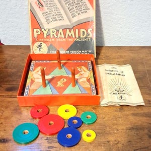 May include: A vintage Pyramids board game featuring an orange box with an illustrated lid, three wooden pegs on a game board, and a set of colorful red, yellow, blue, and green circular disks. Includes a paper instruction booklet titled The Solution of Pyramids.