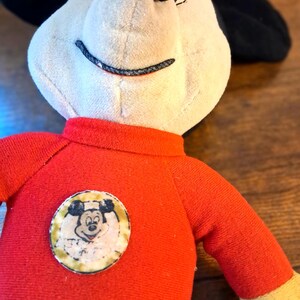 K&ouml;nnte beinhalten: Ein Vintage-Mickey-Mouse-Pl&uuml;schtier. Das Pl&uuml;schtier hat ein cremefarbenes Gesicht, schwarze Ohren und ein rotes Hemd mit einem kreisf&ouml;rmigen Aufn&auml;her mit Mickeys Gesicht. Die Arme sind rot und gelb. Das Spielzeug befindet sich auf einer Holzoberfl&auml;che.