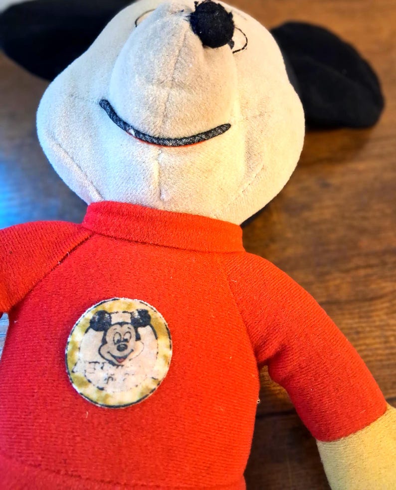 K&ouml;nnte beinhalten: Ein Vintage-Mickey-Mouse-Pl&uuml;schtier. Das Pl&uuml;schtier hat ein cremefarbenes Gesicht, schwarze Ohren und ein rotes Hemd mit einem kreisf&ouml;rmigen Aufn&auml;her mit Mickeys Gesicht. Die Arme sind rot und gelb. Das Spielzeug befindet sich auf einer Holzoberfl&auml;che.