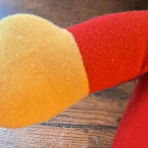 K&ouml;nnte beinhalten: Nahaufnahme eines Pl&uuml;schtierarms. Der Arm ist haupts&auml;chlich rot mit einer gelben Hand. Der Stoff wirkt weich und flauschig. Das Design des Spielzeugs ist einfach und farbenfroh und deutet auf einen Kinderartikel hin.