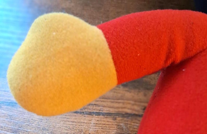 K&ouml;nnte beinhalten: Nahaufnahme eines Pl&uuml;schtierarms. Der Arm ist haupts&auml;chlich rot mit einer gelben Hand. Der Stoff wirkt weich und flauschig. Das Design des Spielzeugs ist einfach und farbenfroh und deutet auf einen Kinderartikel hin.