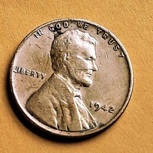 May include: A silver-toned United States one-cent coin featuring the profile of Abraham Lincoln facing right. The text IN GOD WE TRUST is arched at the top, LIBERTY is on the left, and the year 1942 is printed on the lower right side.