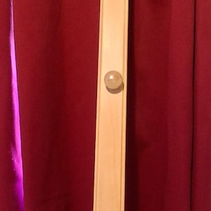 May include: A light-colored wooden coat rack with three round knobs. The rack is rectangular and stands vertically against a dark red curtain. The knobs are evenly spaced along the rack.